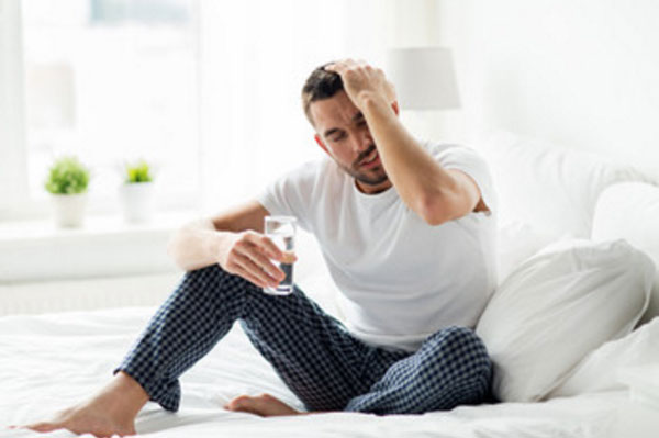 Мужчина сидит на кровати со стаканом воды и держится за голову