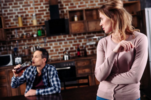 Мужчина пьет с бутылки, сидя за столом. Жена на него смотрит