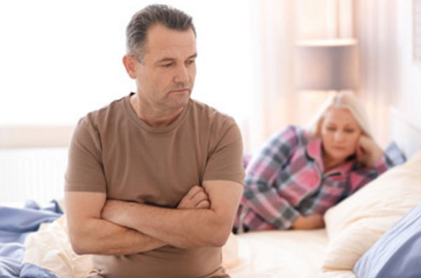 Мужчина сидит на кровати со сложенными руками. Он очень расстроенный. Позади него лежит жена