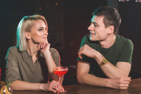Парень говорит что-то девушке, которая сидит рядом за барной стойкой. У нее бокал с алкогольным напитком