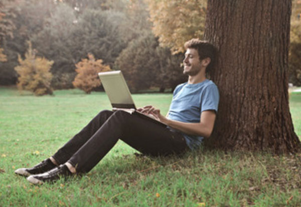 Парень сидит под деревом и что-то печатает на ноутбуке