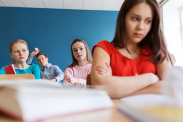 Расстроенная девочка сидит на передней парте. На задней на нее с осуждением смотрят одноклассницы. В конце класса мальчик собирается кинуть в нее скомканный лист бумаги