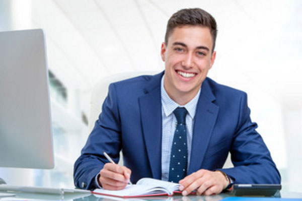 Улыбчивый парень сидит перед компьютером, что-то записывает в ежедневник