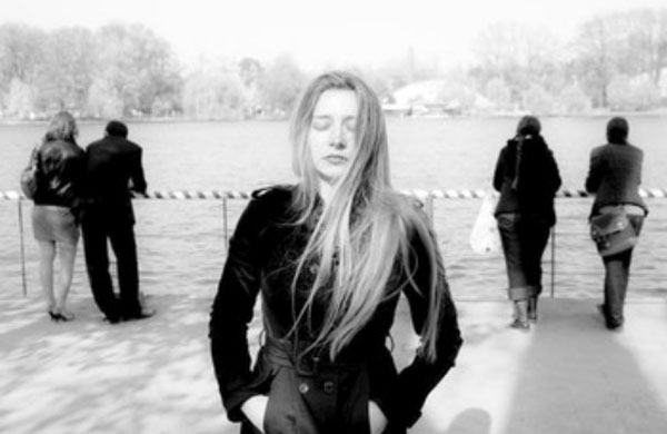 Женщина с закрытыми глазами стоит в задумчивости. Сзади нее остались люди, которые смотрят в другую сторону