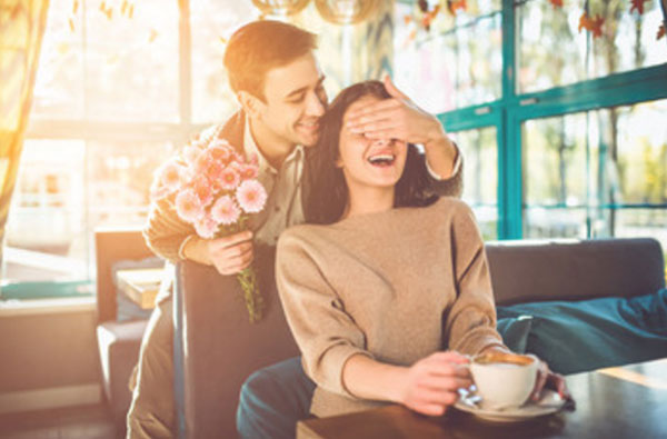 Девушка сидит в кафе с чашкой чая. Сзади подкрался парень с букетом цветов