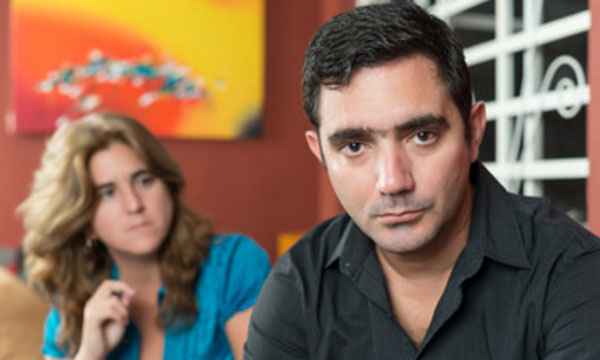 Мужчина смотрит прямо. На заднем плане сидит женщина, которая на него смотрит