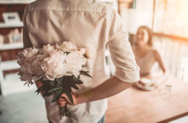 Парень подошел к столу, за котором сидит девушка. За спиной он прячет букет