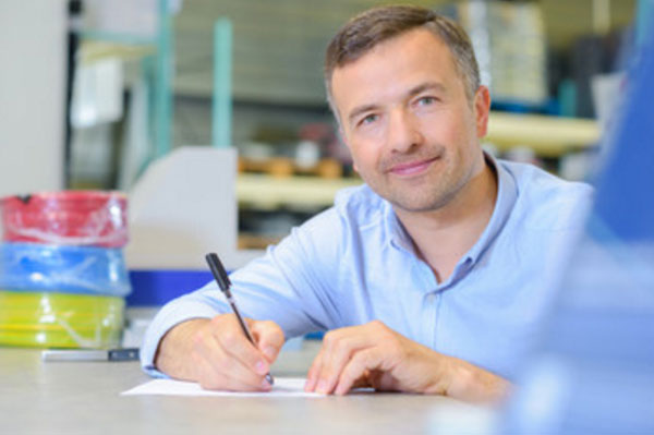 Мужчина что-то пишет на листке бумаги. Смотрит в камеру