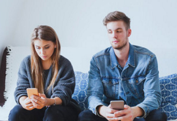 Парень с девушкой сидят на диване, у обоих в руках телефоны. Девушка смотрит в свой экран, а парень аккуратно подглядывает