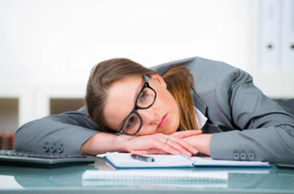 Девушка в костюме и очках лежит на рабочем столе. У нее грустный взгляд