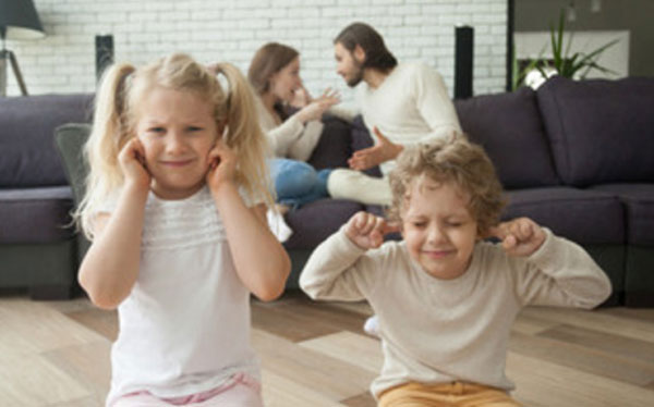 Дети сидят на полу, закрыв уши. На заднем плане родители ругаются, сидя на диване