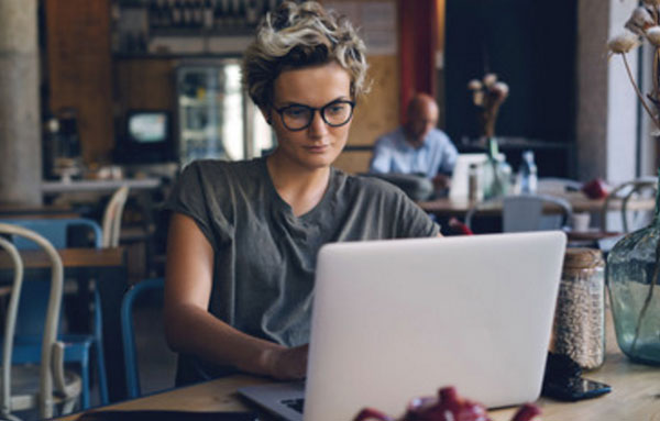 Женщина в очках, простой серой футболку, растрепаной прической и без макияжа печатает на ноутбуке