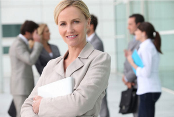 Женщина в костюме улыбается. В руках у нее доклад, сзади стоят люди