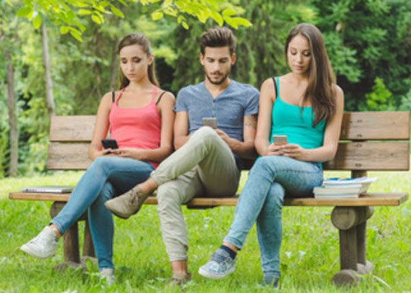Две девушки и парень сидят в одинаковой позе на скамье и клацают в телефонах