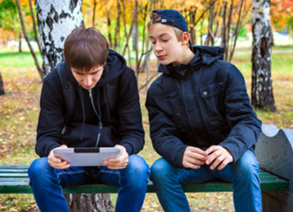 Мальчик смотрит на гаджет парня, сидящего рядом на скамейке