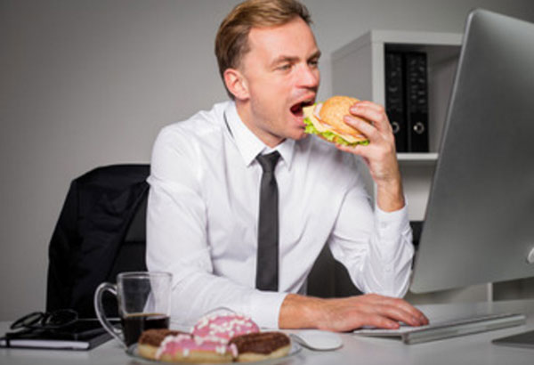 Мужчина сидит за ноутбуком и ест бургер. На столе лежат пончики