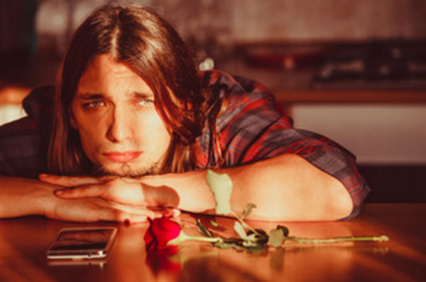 Грустный парень сидит за столом, опустил голову. На столе лежит роза и телефон