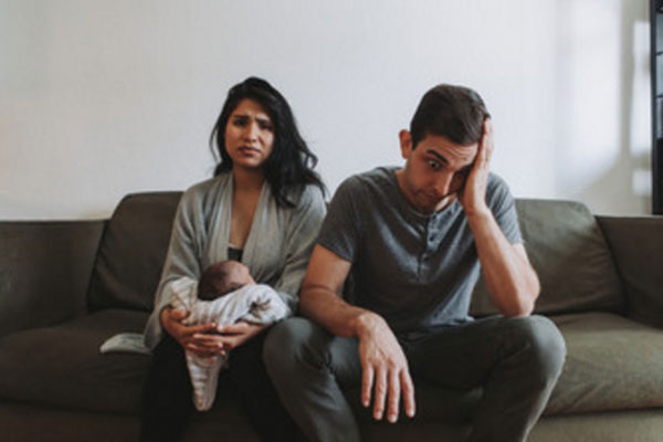 На диване сидит расстроенная женщина с грудным ребенком и мужчина, который в шоке от ситуации