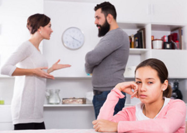 Девочка в расстроенных чувствах сидит за столом. На заднем плане ее родители ссорятся