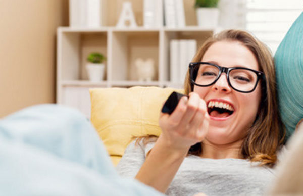 Девушка в очках лежит на диване, держит пульт в руке и смеется