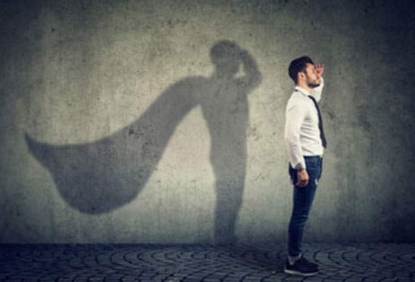 Мужчина стоит, держа руку у виска. Сзади него тень на стене, у которой за спиной расправляется плащ супергероя