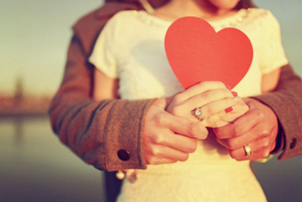 Мужчина сзади обнимает девушку, держит ее руки в своих, она держит картонное сердце