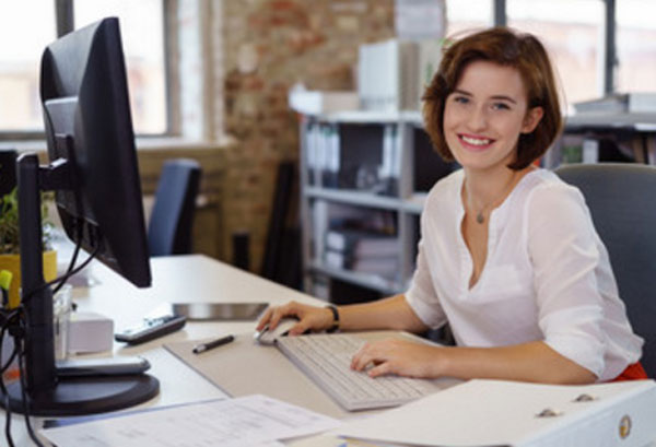 Женщина сидит за рабочим столом, перед ней компьютер, на столе бумаги, она улыбается