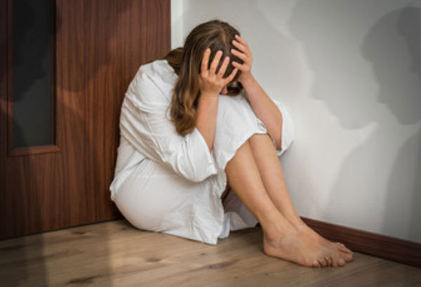 Женщина сидит на полу и держится за голову, пряча лицо. Вокруг нее тени людей