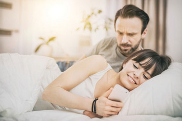 Муж и жена в кровати. Она смотрит в телефон и улыбается. Мужчина насторожен, пытается увидеть, что там происходит