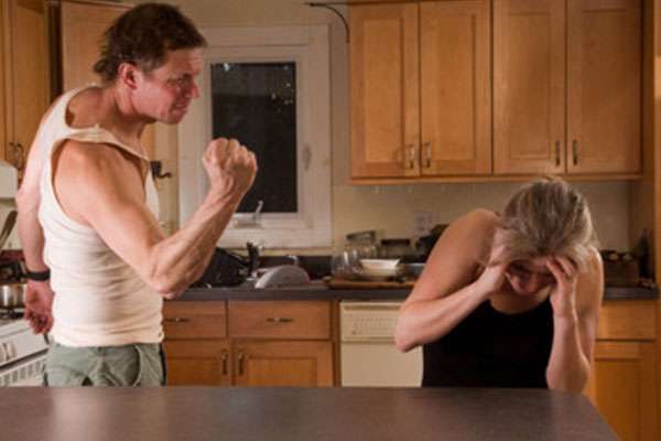 Женщина сидит за столом, испугана, пытается спрятаться от удара. Мужчина угрожает ей кулаком