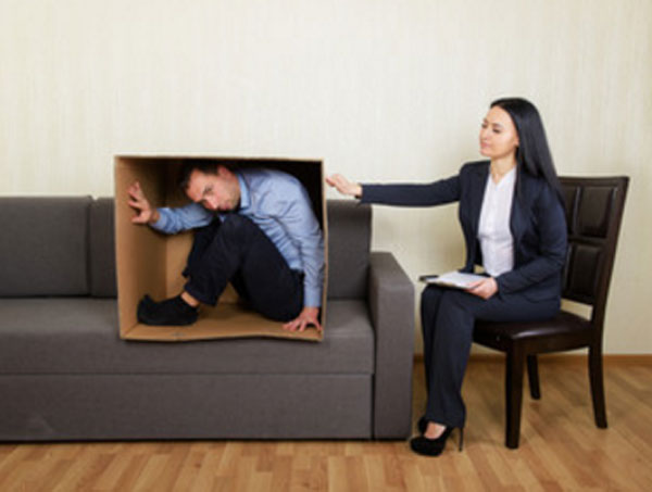 Мужчина сидит в коробке, которая стоит на диване. Рядом на стуле сидит психолог