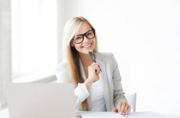 Девушка в очках и костюме сидит перед ноутбуком и улыбается