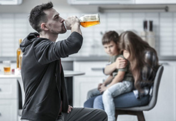 Мужчина бьет с бутылки. На заднем фоне его жена с ребенком на руках