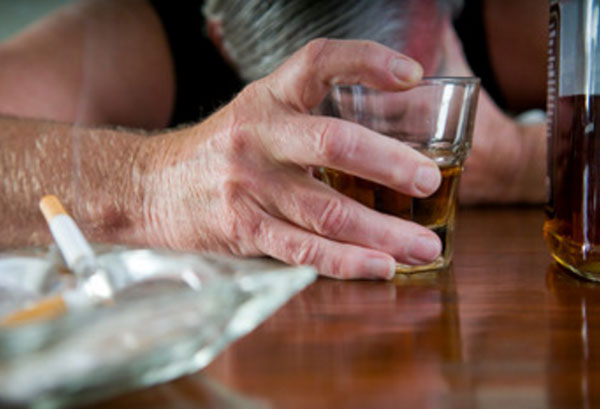 Мужчина опустил голову на стол. В руке у него рюмка с алкоголем. На столе лежат окурки