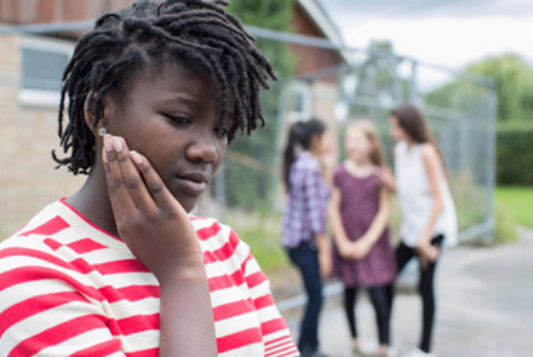 Девушка афроамериканка стоит одна. Она расстроена. На заднем плане судачат девчонки с белым цветом кожи