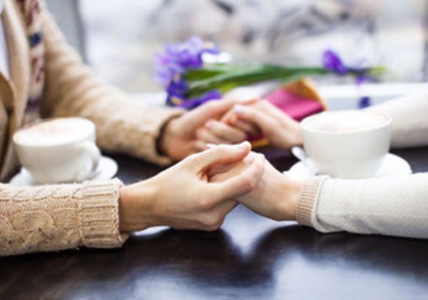 Руки девушки и парня. Пара сидит за столом напротив друг друга, на котором стоят чашечки и держатся за руки